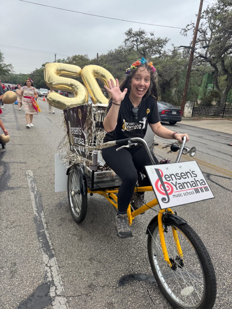 Woman riding a yellow decorated tricycle in a parade, waving at the camera; large gold '50' balloons and banners for Lensen's Yamaha music school are attached to the vehicle.